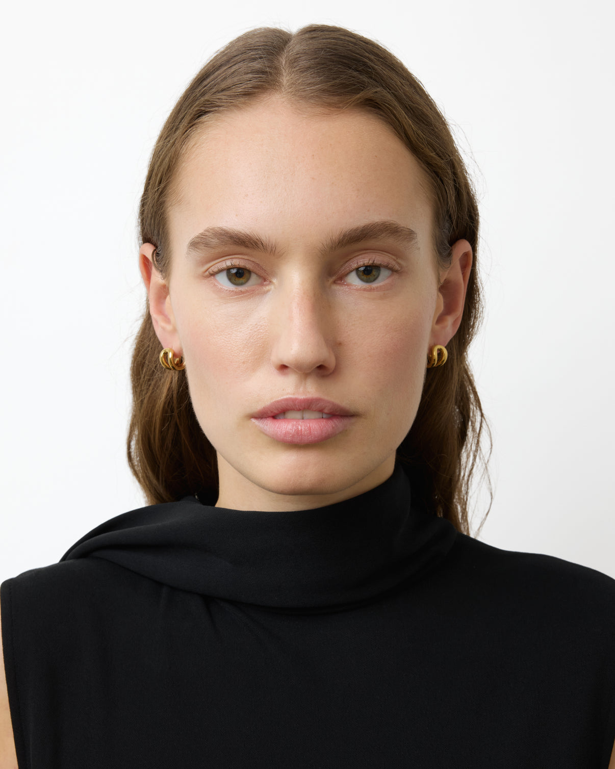 A woman with light skin and long brown hair wears a black sleeveless top and Lie Studio Asta Earrings, facing forward against a plain white background.