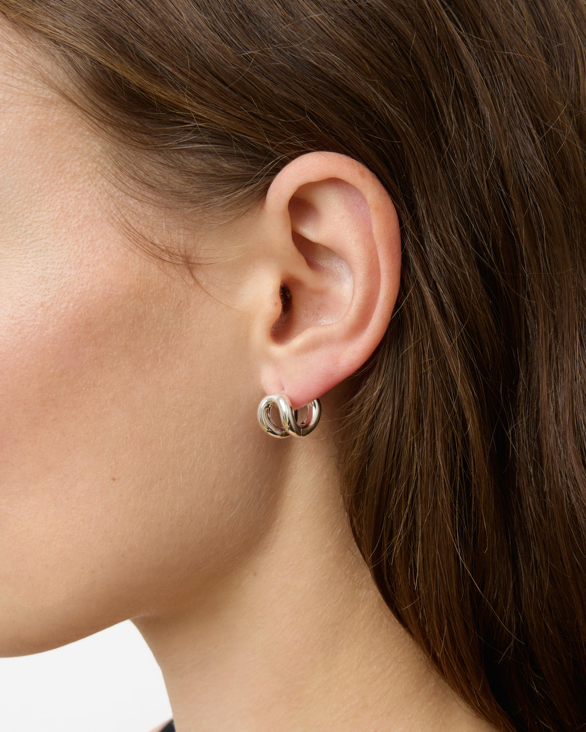 Close-up of a woman's ear with long brown hair, wearing a small, shiny sterling silver hoop Asta Earring from Lie Studio, highlighting its refined silhouette.