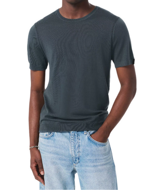 A person wearing the Rag & Bone Aulton Knit Tee, a dark fine jersey crewneck, paired with light blue jeans and one hand in their pocket, stands against a plain white background.