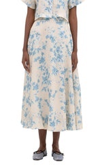 A person elegantly poses against a white backdrop wearing a cream dress with blue floral patterns and the Agua by Aguabendita Bergamota Skirt, paired with gray ballet flats.