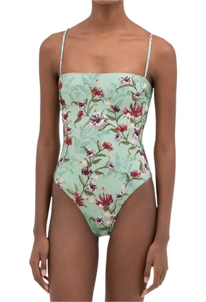A person wears the Durazno Embroidered One Piece by Agua By Aguabendita, featuring a floral pattern and thin shoulder straps, made from recycled polyester, standing against a white background.