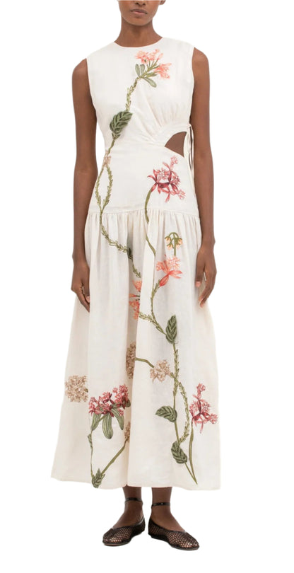 Person wearing an Agua By Aguabendita Otono Midi Dress, a sleeveless white linen piece with floral embroidery and side cut-out details, paired with brown woven shoes.