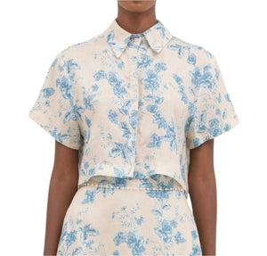 Person wearing an Agua by Aguabendita Virtuosa Cropped Shirt, featuring a blue Bougainvillea floral print on a beige pure linen, short-sleeve button-up.