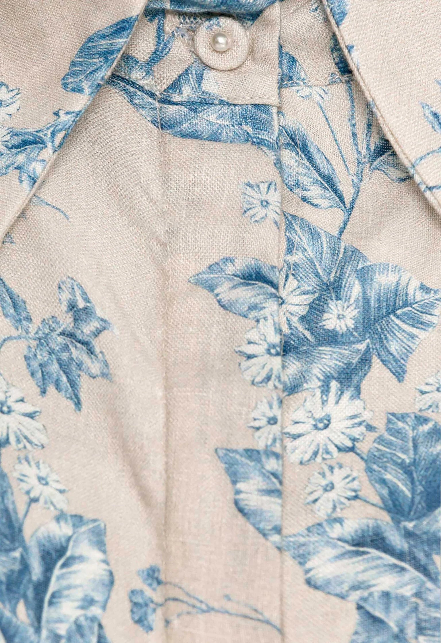 Close-up of the Agua by Aguabendita Virtuosa Cropped Shirt in beige linen with a blue floral print, featuring a partially buttoned collar.