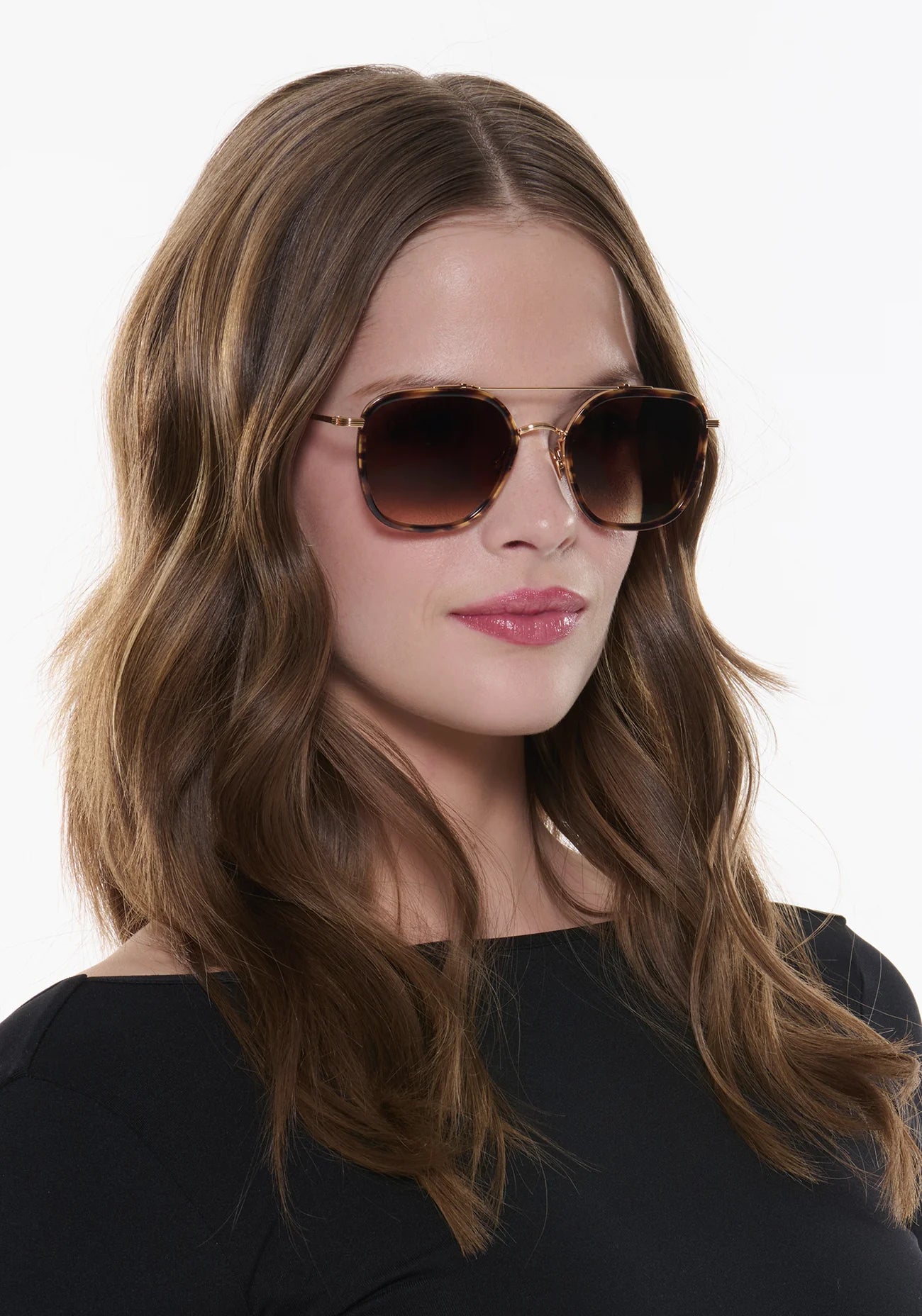 A woman with wavy brown hair wears Krewe Austin sunglasses and a black top, angled slightly to the side against a white background—an effortlessly chic look that complements many face shapes.