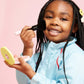 A young girl with braided hair smiles as she applies blush to her cheek using a brush and holds a compact powder case from the Super Smalls Mom's Makeup Play Kit, set against a pink background.
