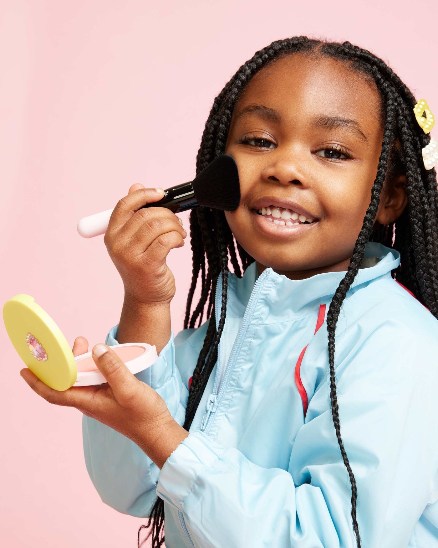 A young girl with braided hair smiles as she applies blush to her cheek using a brush and holds a compact powder case from the Super Smalls Mom's Makeup Play Kit, set against a pink background.