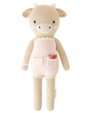 Ava the Cow Powder Pink by Cuddle + Kind is a 20" knitted doll in a pink apron with pockets, one holding a strawberry—perfect for birthdays or adding a cozy touch of the meadow to your home.