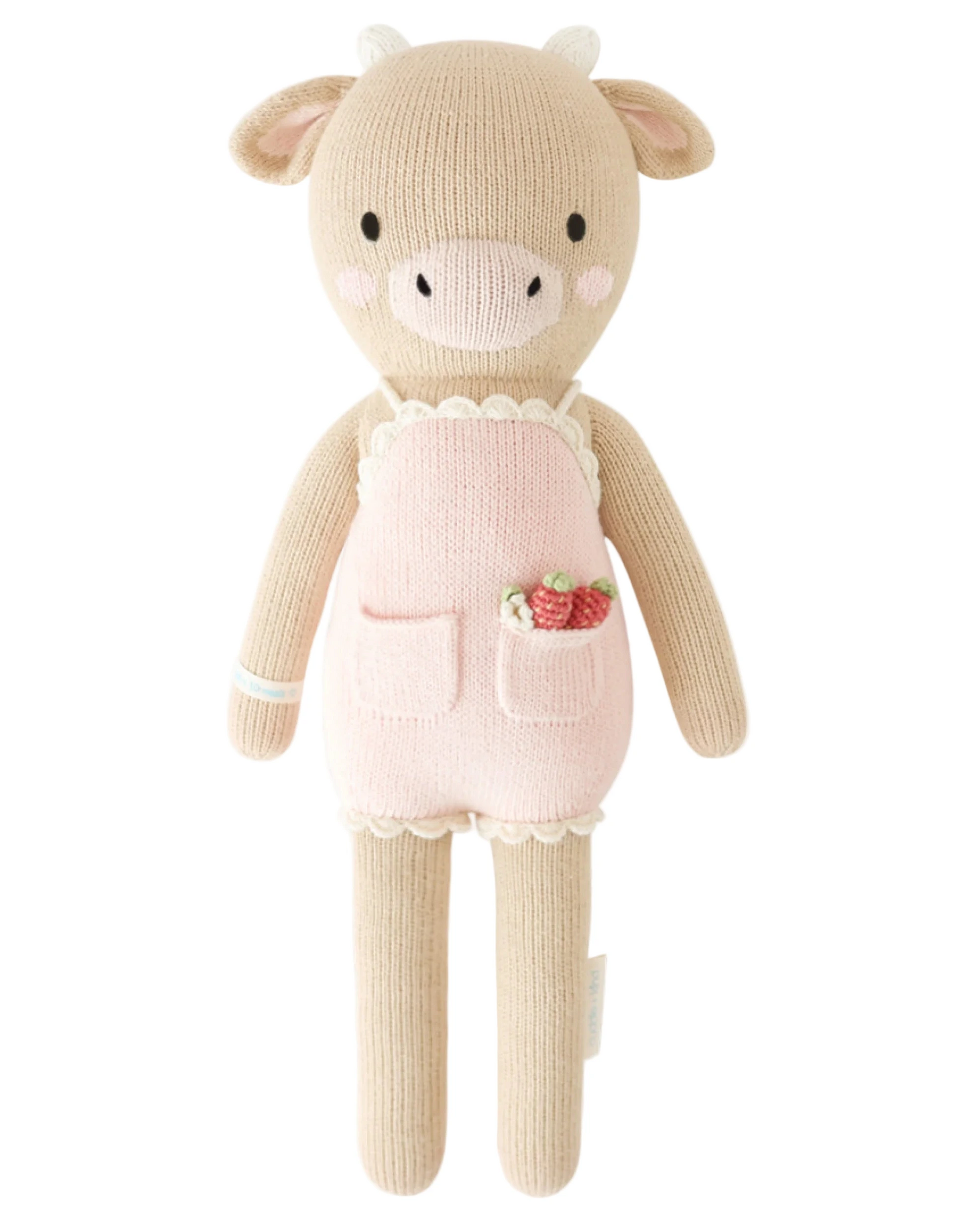 Ava the Cow Powder Pink - Little 13" by Cuddle + Kind is a knitted plush toy with a beige body, white horns, and a powder pink apron with pockets—one holding small strawberries—making it a charming birthday gift or friendship symbol.