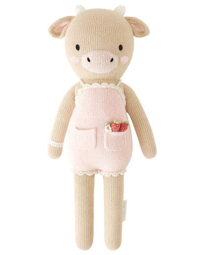 Ava the Cow Powder Pink - Little 13" by Cuddle + Kind is a knitted plush toy with a beige body, white horns, and a powder pink apron with pockets—one holding small strawberries—making it a charming birthday gift or friendship symbol.