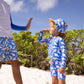 Two children, each wearing Snapper Rock's Shark Squad Reversible Bucket Hat and matching swimsuits, stand on a sandy beach with greenery behind them, giving each other a high five.