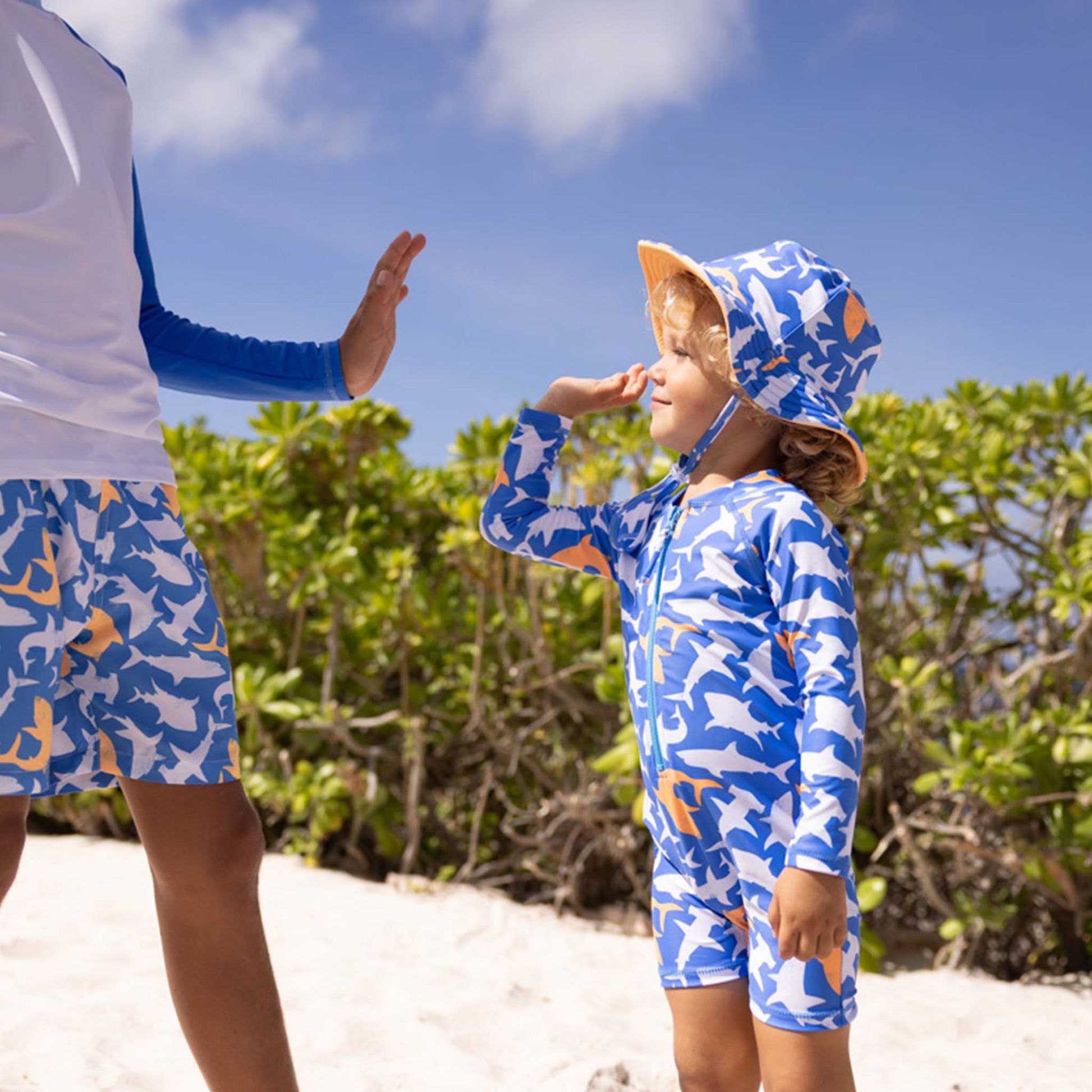 Two children, each wearing Snapper Rock's Shark Squad Reversible Bucket Hat and matching swimsuits, stand on a sandy beach with greenery behind them, giving each other a high five.