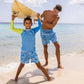 A child stands on a beach with a surfboard overhead, wearing the vibrant Snapper Rock Citron Splash Rashguard swim top. An adult, smiling nearby, is also in blue patterned swim shorts. Both enjoy the ocean backdrop and clear sky, ensuring perfect sun protection.