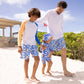Two adults and a child, all in matching blue and white swimwear, walk on a sunny beach. The child wears the Snapper Rock Boys' Blue Papaya LS Rash Top with UPF50+ protection.