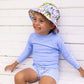 A smiling child wears the Snapper Rock Baby Boys' Swaside Blue LS Let rash top, paired with floral bucket hat and blue gingham swim shorts with an elastic waistband, sitting against a white wooden wall.