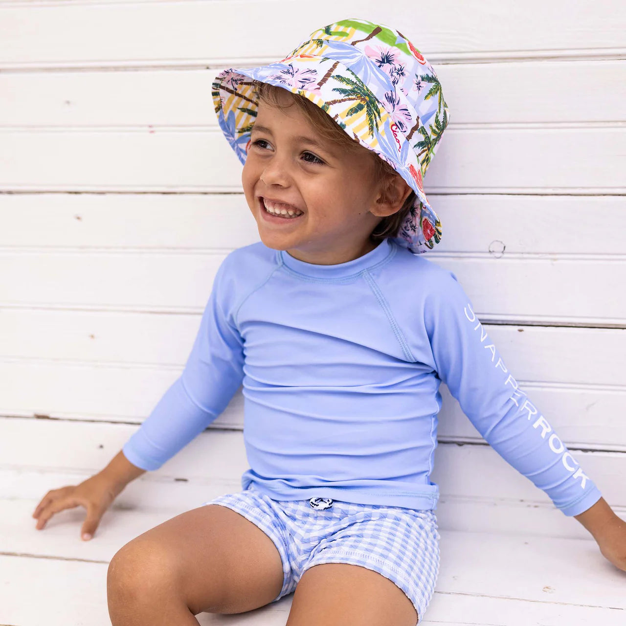 A smiling child wears the Snapper Rock Baby Boys' Swaside Blue LS Let rash top, paired with floral bucket hat and blue gingham swim shorts with an elastic waistband, sitting against a white wooden wall.