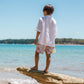 A child stands barefoot on a driftwood log by the water in the Snapper Rock Boys' Frankie White Resort Shirt, with a blue sky and distant trees in the background.