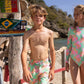 Two children in matching Snapper Rock Coastal Shells Swim Shorts stand near a beach. The shorts boast an adjustable drawcord waist for a perfect fit. A signpost with various destinations is seen in the background, along with trees and a rocky shoreline.