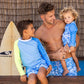 A man and two children, all wearing Snapper Rock Jawesome Waves swim shorts, sit on a wooden bench near a surfboard.