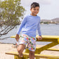 A boy in a light blue long-sleeve shirt and Snapper Rock Boys' Ciao Paradiso Swim Short stands on a yellow picnic table by the water, with sand and a tree in the background.
