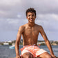 A young man sits on a dock by the water, smiling, wearing Snapper Rock Boys’ Cali Palms Recycled Swim Shorts with a comfort liner. Boats and a cloudy sky form the background.