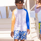 A boy wearing Snapper Rock Boys' Shark Squad Swim Shorts and a long-sleeve color-block rash guard stands outside by a white beach hut.