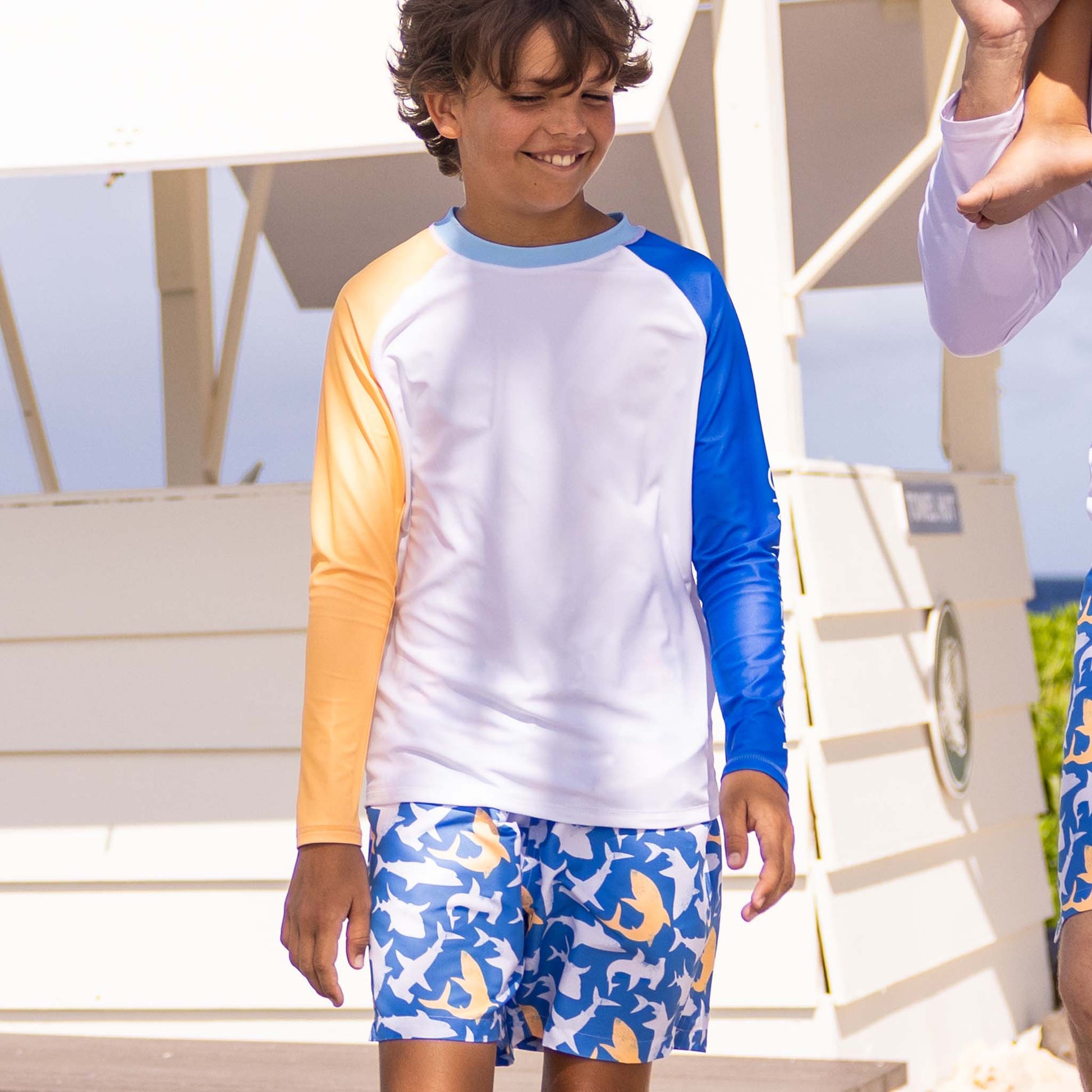 A boy wearing Snapper Rock Boys' Shark Squad Swim Shorts and a long-sleeve color-block rash guard stands outside by a white beach hut.