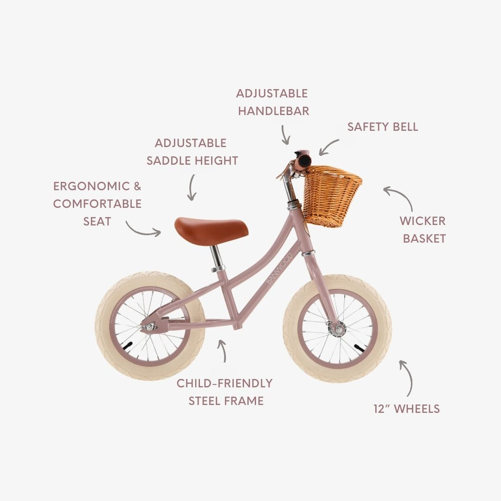 Kids’ Vintage First Go Balance Bike by Banwood in pink, featuring white 12″ wheels, a classic wicker basket, brown ergonomic seat, safety bell, adjustable handlebar and saddle, and a sturdy steel frame. Labels highlight each feature of this stylish children’s bike.