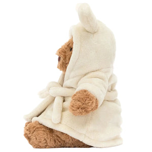 The Jellycat Bartholomew Bear Bathrobe plush sits upright in profile on a white background, wearing a cream-colored hooded robe with cute ears.