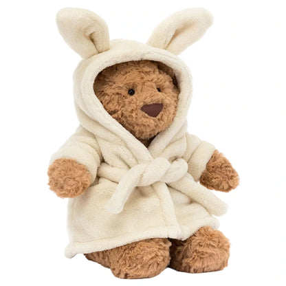 Bartholomew Bear Bathrobe by Jellycat sits upright, wearing a cream hooded robe with bunny ears and a tied belt—this adorable plush bear is perfect for cozy hugs.