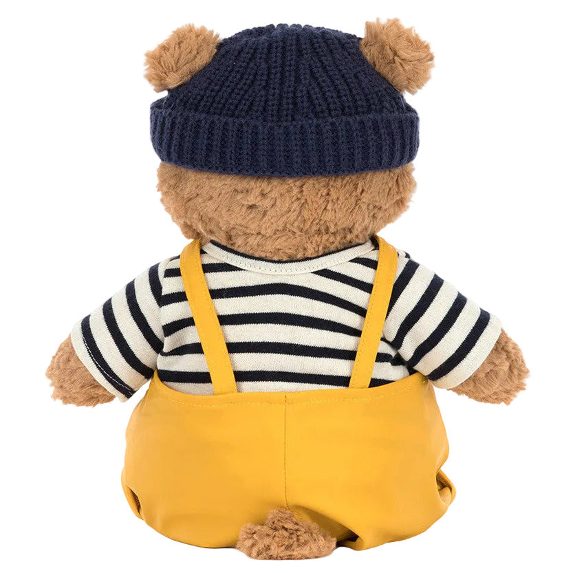 The Jellycat Bartholomew Bear Seafarer Outfit features a plush teddy bear facing away, dressed in a navy knit hat, striped shirt, and yellow strapped overalls—perfectly ready for a boat trip adventure.