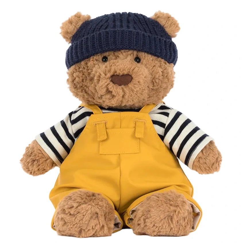 Bartholomew Bear Seafarer Outfit by Jellycat features a plush bear in a navy knit hat, black and white striped shirt, and yellow overalls, sitting against a white background, ready for seaside adventures.