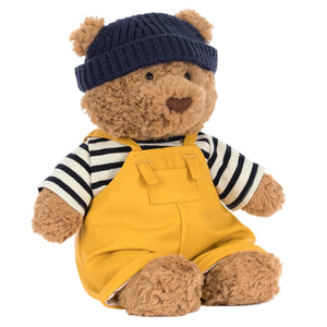 Jellycat’s Bartholomew Bear Seafarer Outfit features a brown bear in a navy knit hat, striped shirt, and yellow overalls, sitting upright against a white background as if ready for an adventure.