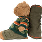 A Jellycat Bartholomew Bear in a Hiking Outfit sits upright, leaning against a rolled-up green sleeping bag.
