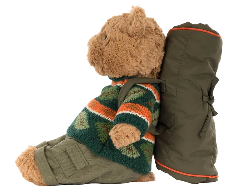 A Jellycat Bartholomew Bear in a Hiking Outfit sits upright, leaning against a rolled-up green sleeping bag.