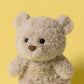 A Jellycat Bartholomew Bear Junior, featuring a fluffy light brown coat, black eyes, and a brown nose, is displayed against a solid yellow background.