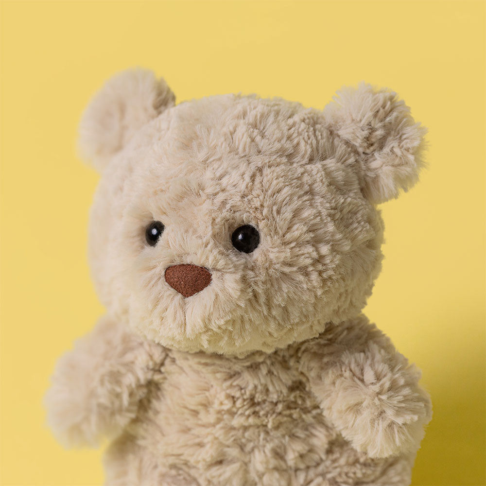 A Jellycat Bartholomew Bear Junior, featuring a fluffy light brown coat, black eyes, and a brown nose, is displayed against a solid yellow background.