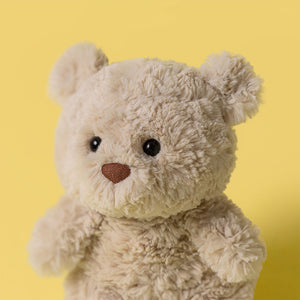 A Jellycat Bartholomew Bear Junior, featuring a fluffy light brown coat, black eyes, and a brown nose, is displayed against a solid yellow background.