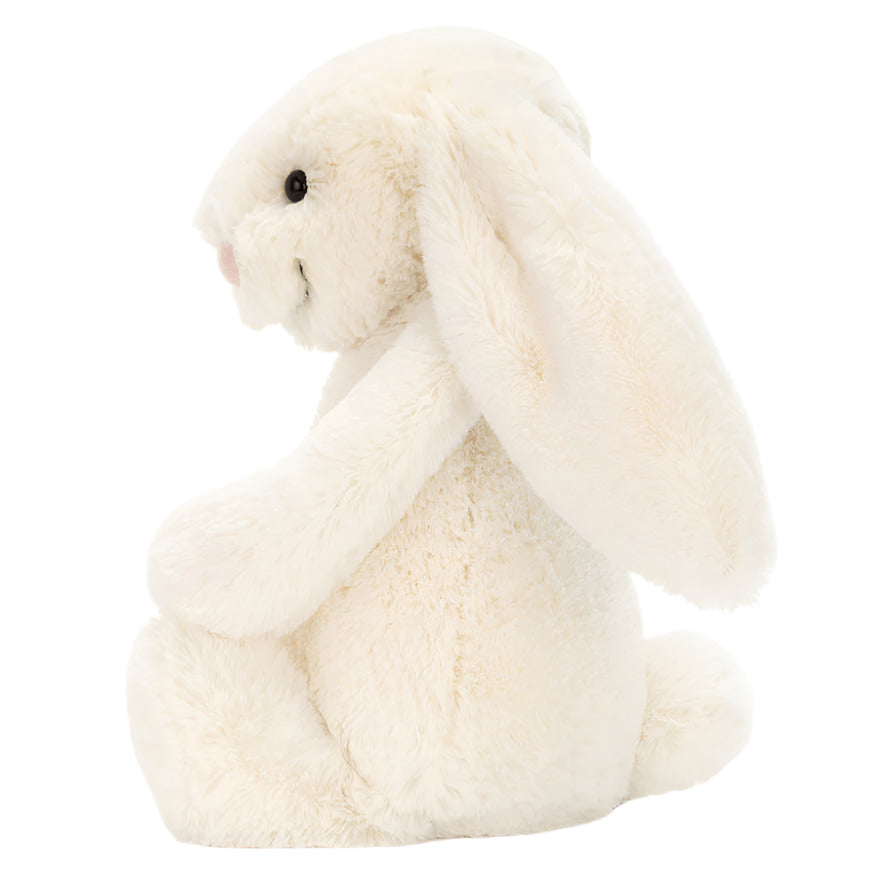 The Jellycat Bashful Bunny Cream Medium sits upright, facing left, with long floppy ears and ultra-soft fur—an ideal cuddly companion for lazy summer days.
