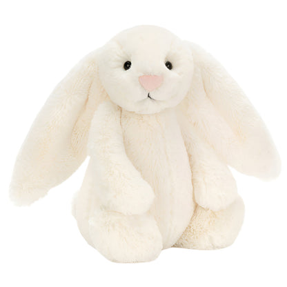 The Jellycat Bashful Bunny Cream Medium, with long floppy ears, black eyes, and a pink nose, sits upright against a white background—perfect for cuddling on relaxed summer days.