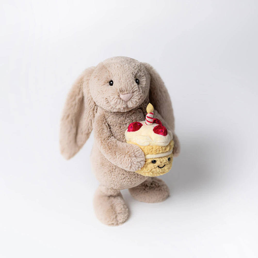 The Jellycat Bashful Beige Bunny 'Birthday' plush holds a cupcake with strawberries, cream, and a lit candle—an adorable birthday gift, shown on a plain white background.