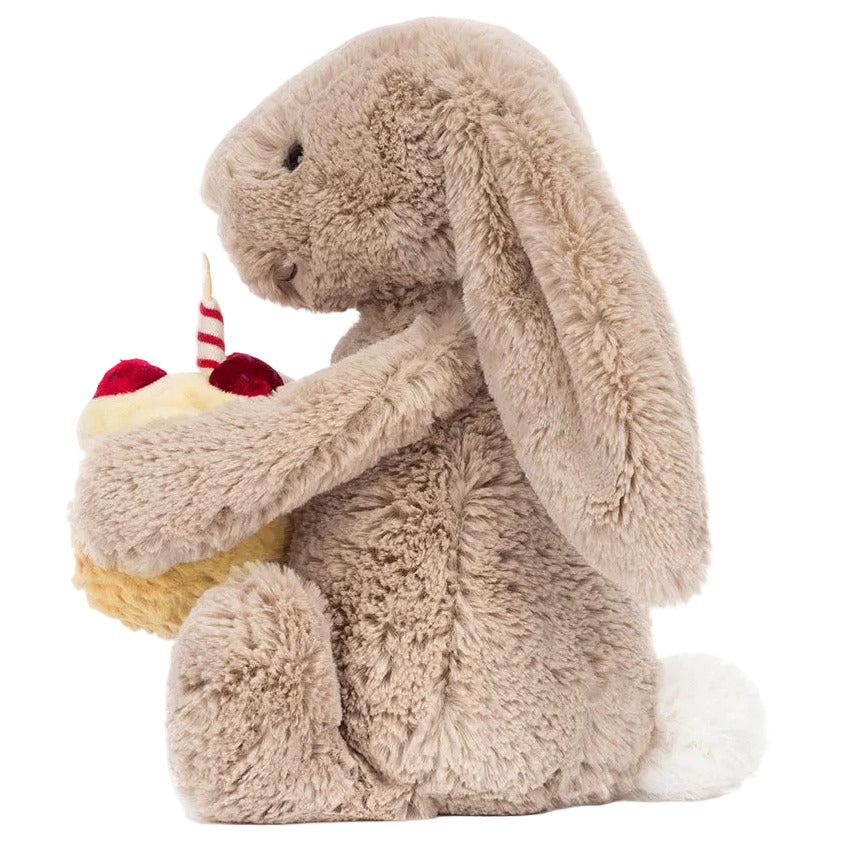 The Bashful Beige Bunny 'Birthday' by Jellycat sits upright with floppy ears, holding a toy cupcake topped with a candle and two red cherries—making it an adorable birthday gift for any child.