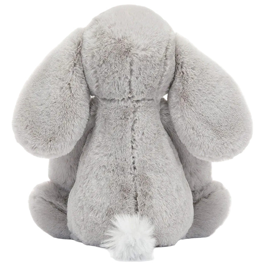 A plush grey Bashful Luxe Elephant Thuddeus from Jellycat sits facing away, highlighting its floppy ears and fluffy tail against a plain background.