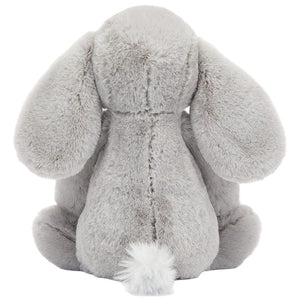 A plush grey Bashful Luxe Elephant Thuddeus from Jellycat sits facing away, highlighting its floppy ears and fluffy tail against a plain background.