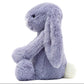 The Bashful Viola Bunny by Jellycat is a soft, plush toy with light purple fur, long floppy ears, black eyes, and a fluffy white tail, shown in side profile.