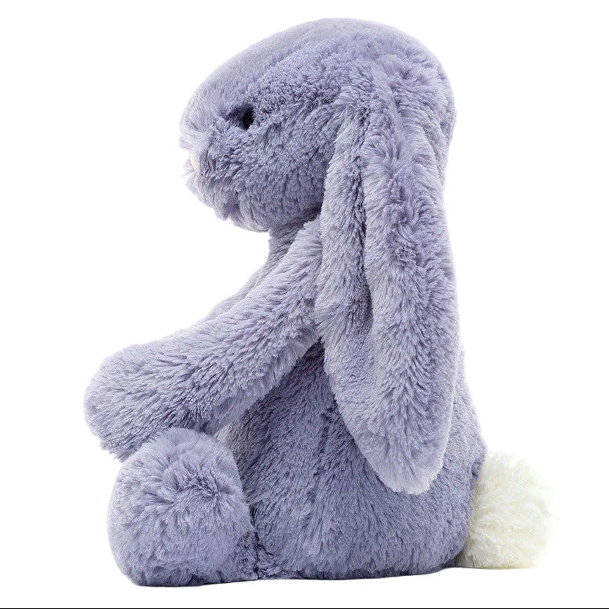 The Bashful Viola Bunny by Jellycat is a soft, plush toy with light purple fur, long floppy ears, black eyes, and a fluffy white tail, shown in side profile.