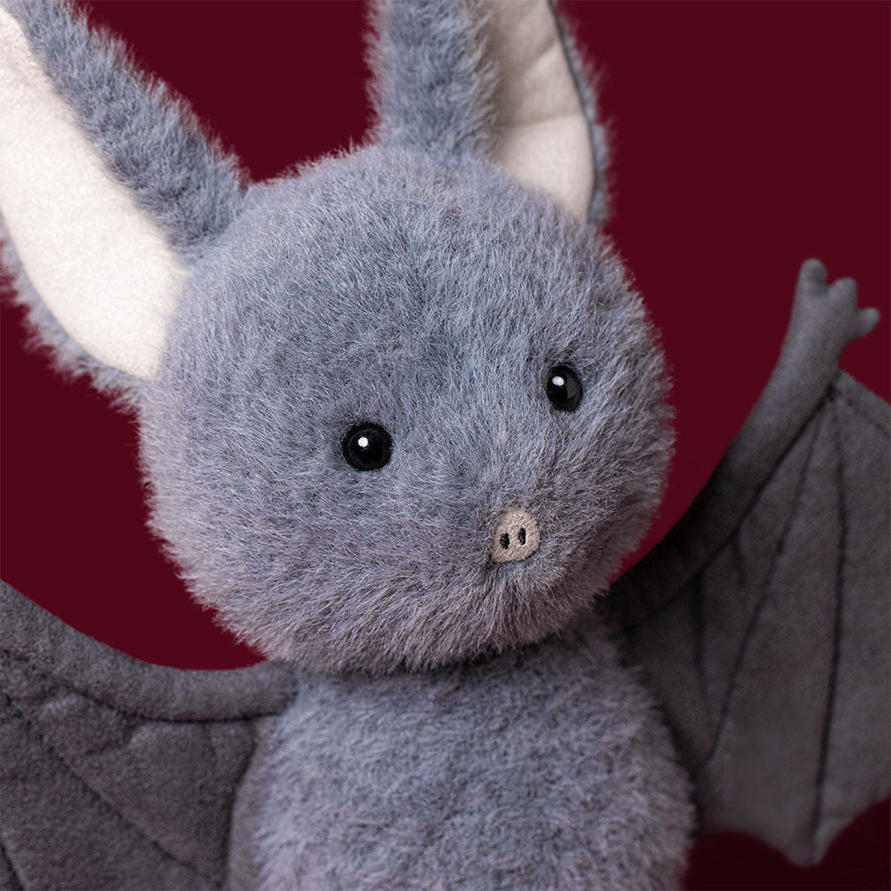 A Jellycat Broox Bat plush toy with gray fur, large white inner ears, black eyes, and fabric wings is displayed against a solid dark red background.