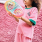 A young child with curly hair stands in front of a pink wall, dressed in a casual pink outfit with green trim and wearing the Bonton Girls’ Livia T-Shirt made from 100% cotton, while holding a colorful plastic spring toy.