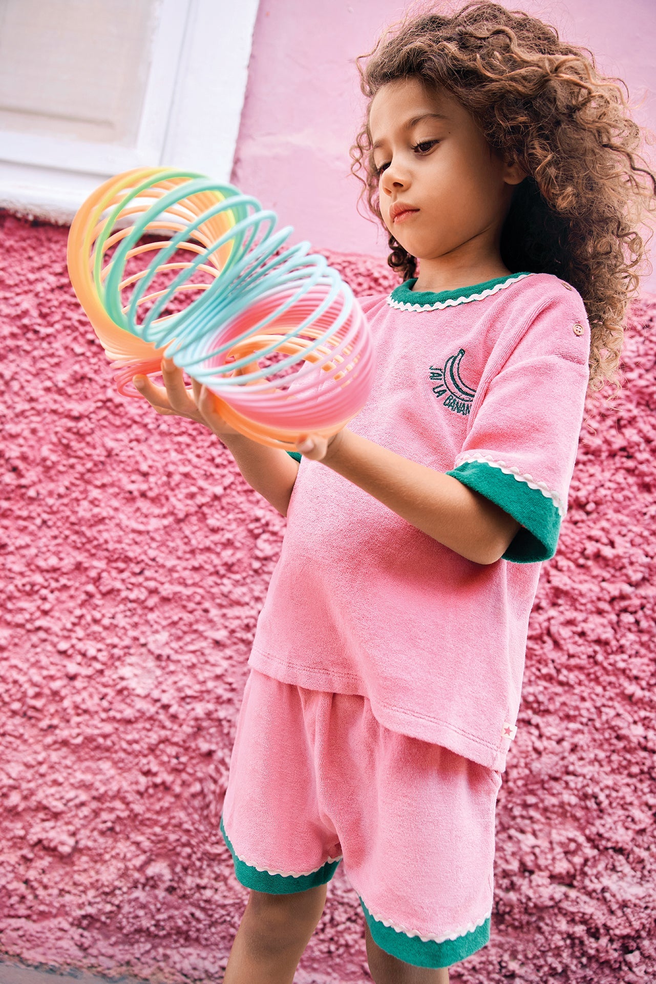 A young child with curly hair stands in front of a pink wall, dressed in a casual pink outfit with green trim and wearing the Bonton Girls’ Livia T-Shirt made from 100% cotton, while holding a colorful plastic spring toy.