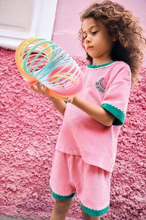 A young child with curly hair stands in front of a pink wall, dressed in a casual pink outfit with green trim and wearing the Bonton Girls’ Livia T-Shirt made from 100% cotton, while holding a colorful plastic spring toy.